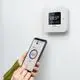 Ein weißer digitaler Thermostat von Theben an der Wand, mit einem verbundenen Smartphone, das die Temperatur und Einstellungen anzeigt. Das Display zeigt aktuelle Werte an.